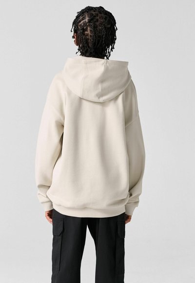 Personne aux cheveux tressés portant un sweat à capuche crème surdimensionné et un pantalon cargo noir, vue de dos sur un fond uni.