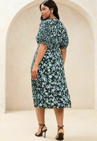 Schwarzes Blumenkleid mit kurzen Puffärmeln, geraffter Taille und knielangem Rock mit blauen und grünen Blumenmustern, kombiniert mit schwarzen Absätzen.