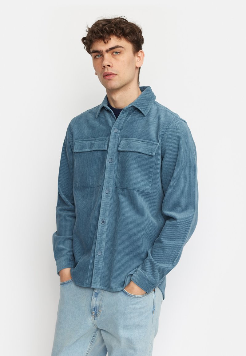 Camicia in corduroy blu con colletto, due tasche sul petto e chiusura a bottoni. Le maniche lunghe presentano polsini con bottoni. Tessuto testurizzato.