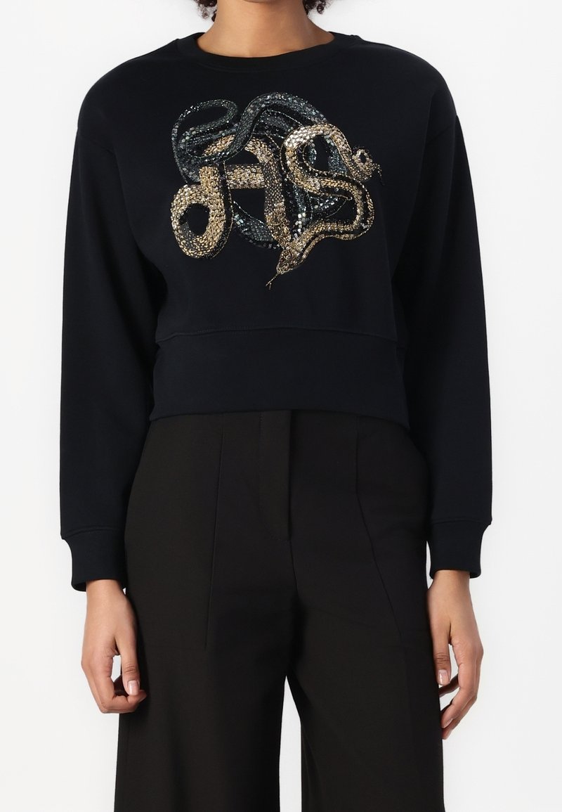 Haut noir à manches longues avec un grand serpent en sequins noirs et dorés, associé à un pantalon noir taille haute.