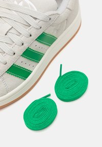 Baskets en suede gris clair avec un design à trois bandes vertes, lacets blancs et semelle en caoutchouc marron. Deux lacets ronds verts sont à côté de la chaussure.