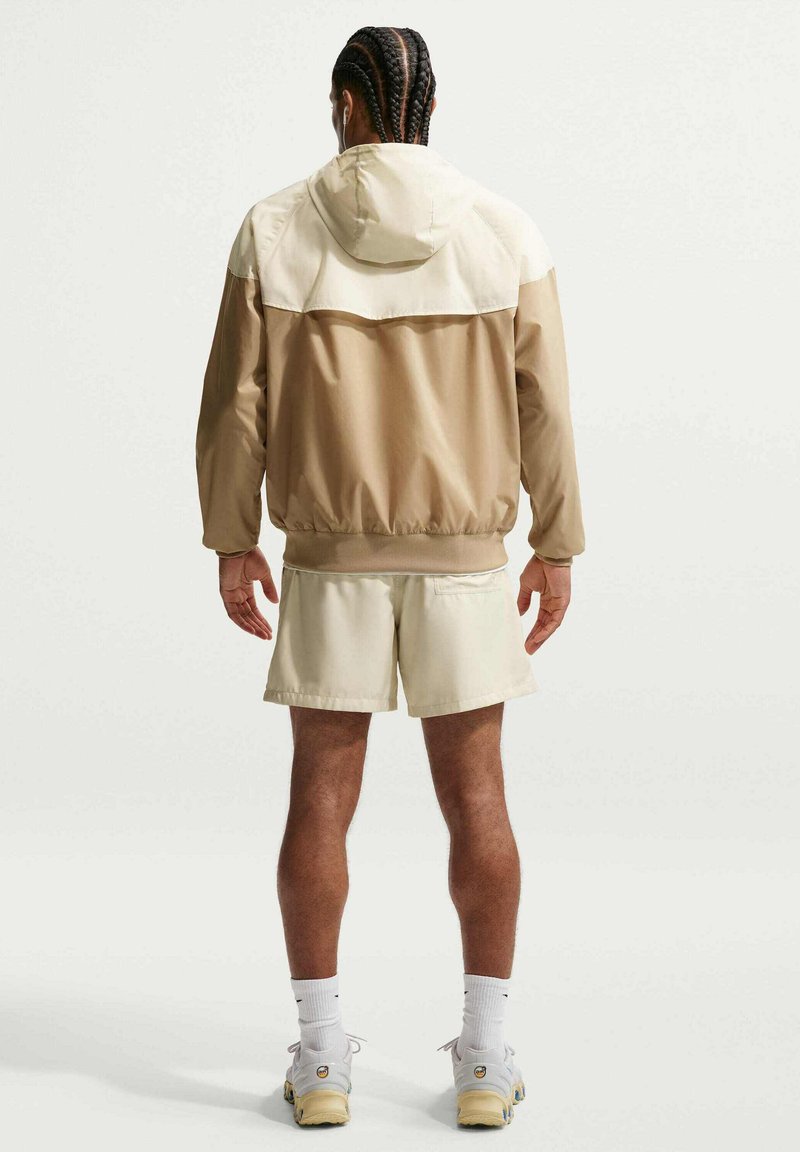 Man met gevlochten haar die een beige en crèmekleurig capuchonjack, crèmekleurige korte broek, witte sokken en witte sneakers draagt en met de rug naar voren staat.