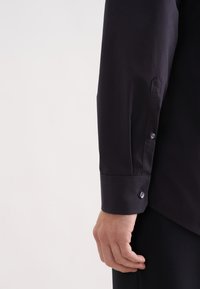 Czarna koszula z długim rękawem o gładkiej fakturze. Posiada mankiet z guzikiem i dopasowany krój, na zdjęciu widoczny jest tylko rękaw i część ramienia.