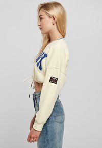 Cropped beige sweatshirt with blue lettering, drawstring hem, and logo patch on sleeve. Worn with high-waisted blue jeans.