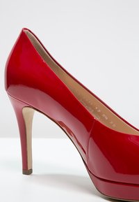 Chaussure à talon haut rouge brillant pour femme, à bout pointu et doublure intérieure beige, posée sur une surface blanche avec un fond neutre.