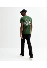 Grön bomulls t-shirt med en vit blommönsterdesign på ryggen. Tillsammans med svarta byxor och svarta sneakers, bärs med en svart keps.