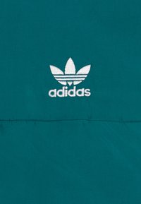 Tīrās audums ar izteiksmīgu balto Adidas trīszaru logotipu un „adidas” tekstu zem tā; gluda tekstūra, minimālists dizains.