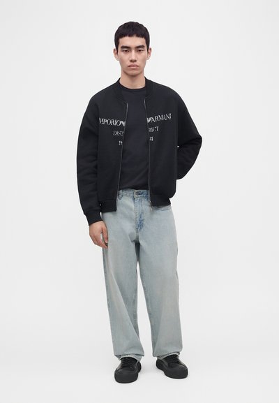 Jeune homme portant une veste zippée noire Emporio Armani, un t-shirt noir, un jean large bleu clair et des baskets noires, debout avec une main dans le dos.