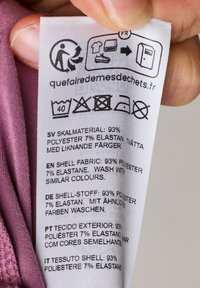 Etykieta z instrukcją pielęgnacji i składem materiałowym: 93% poliester, 7% elastan. Zawiera nadrukowany tekst w kilku językach. Kolor: biały z purpurowym akcentem.