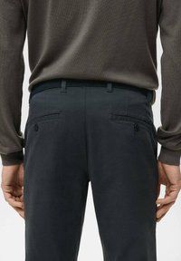 Mężczyzna ubrany w ciemnoszare spodnie z zapinanymi na guziki tylnymi kieszeniami oraz brązowy sweter z długim rękawem włożony do środka, ręce swobodnie opadające wzdłuż ciała.