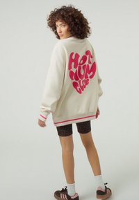 Krém színű pulóver vastag rózsaszín "HOT MUM CLUB" felirattal a hátulján. Rugalmas mandzsettákkal és szegéllyel rendelkezik, fekete leopárdmintás rövidnadrággal és sportcipőkkel párosítva.
