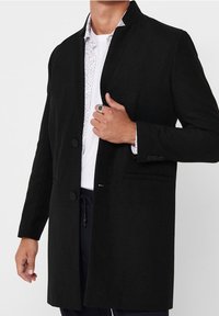 Zwarte wollen jas met een hoge kraag, single-breasted ontwerp, twee knopen en een borstzak, gedragen over een wit shirt met een patroon.