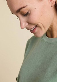 Sweatshirt en coton vert avec un col rond, texture douce et détails cousus, porté par une personne à la peau claire.