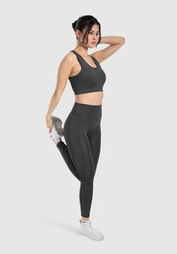 Sujetador deportivo negro y leggings de cintura alta hechos de un tejido suave y elástico. La modelo está estirándose, mostrando un diseño ajustado y contorneado.