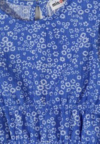 Tissu bleu à surface texturée présentant un petit imprimé floral blanc et une taille froncée avec une étiquette blanche cousue à l'encolure.