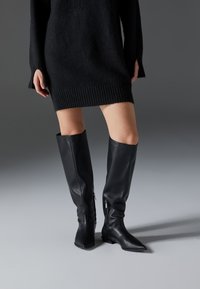 Schwarze Lederstiefel über dem Knie mit spitzen Zehen, flachen Sohlen und seitlichen Reißverschlüssen. Glatte Textur, elegantes Design ohne Verzierungen.