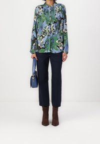 Bloemrijke blouse met lange mouwen in blauw, groen en zwart met een kraag; gecombineerd met marineblauwe broek. Bruine enkellaarsjes en een blauwe handtas.