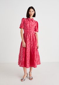 Mujer vistiendo un vestido midi rosa floral con mangas abullonadas y sandalias de tacón alto con tiras plateadas, de pie contra un fondo blanco.