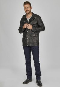 Schwarze Lederjacke mit Druckknöpfen, Reißverschluss-Taschen und Kapuze, kombiniert mit dunklen Jeans und schwarzen Stiefeln, auf einem grauen Hintergrund.