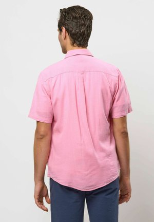 Lühikeste tumedate juustega mees, kandes lahedat roosat lühikeste varrukatega särki ja tumedat sinist püksid, seisab seljaga vaataja poole ühevärvilise tausta ees.