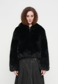 Μαύρο τζάκετ από ψεύτικη γούνα με ψηλό γιακά, κλείσιμο με φερμουάρ και φαρδιά μανίκια, συνδυασμένο με σκουρόχρωμη πλισέ φούστα με λεπτομέρεια ζώνης.