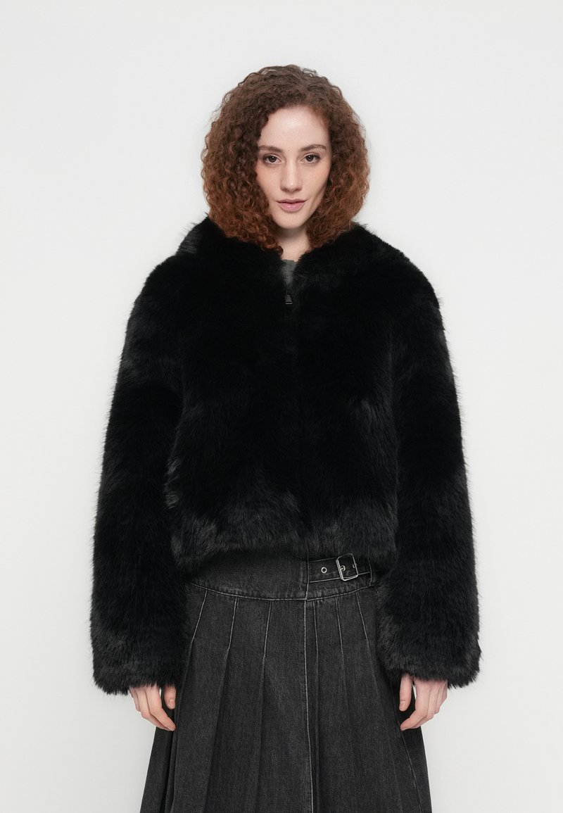 Μαύρο τζάκετ από ψεύτικη γούνα με ψηλό γιακά, κλείσιμο με φερμουάρ και φαρδιά μανίκια, συνδυασμένο με σκουρόχρωμη πλισέ φούστα με λεπτομέρεια ζώνης.