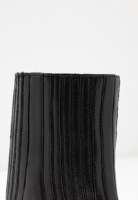 Lähikuva mustastNahkasaapast, jossa on pystysuorat ommellut paneelit ja teksturoitu pinta valkoista taustaa vasten.