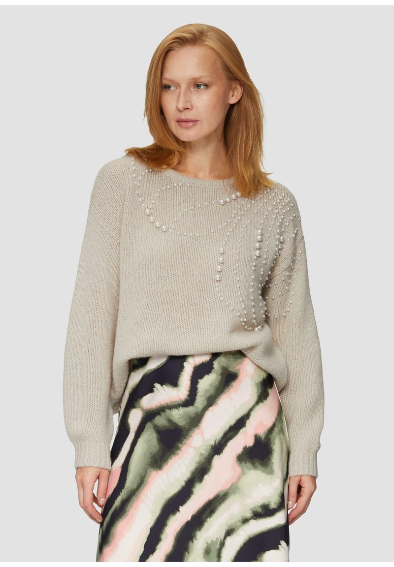 Beige gestrickter Pullover mit Perlenakzenten in einem spiralförmigen Muster; kombiniert mit einem mehrfarbigen Rock, der geschwungene grüne und rosa Designs zeigt.