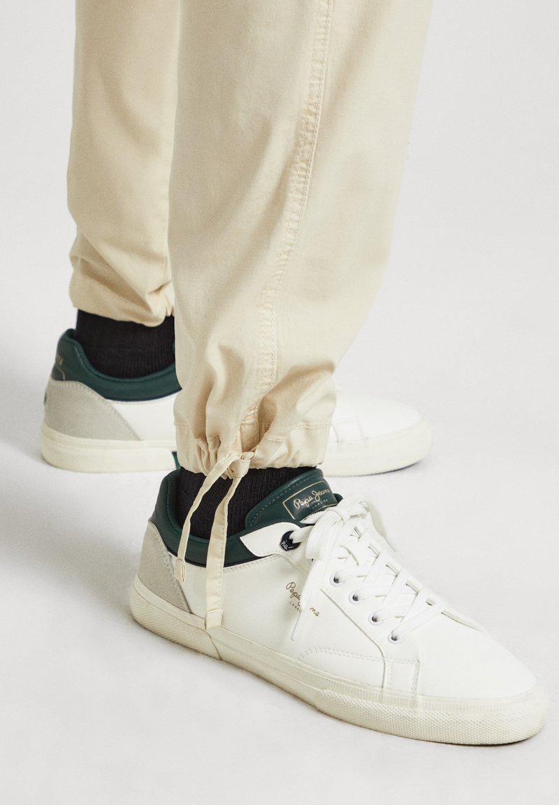 Zapatillas blancas con acentos verdes, suela de goma texturizada y detalle de logotipo. Combinadas con joggers color crema con puños elásticos.