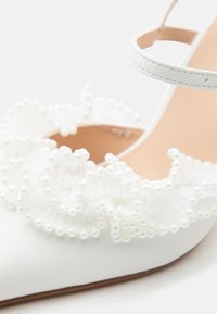 Chaussure blanche à talons hauts avec bride à la cheville et volant décoratif de perles blanches et de tissu le long du bord supérieur.