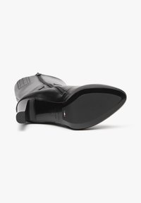 Bottine en cuir noir avec un bout pointu, un talon haut et un détail de fermeture éclair. Texture lisse avec des accents de couture verticale sur le côté.