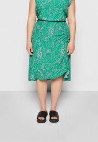 Vestido verde com um padrão floral em contornos rosas, apresentando um detalhe franzido na lateral. Combinado com sandálias pretas de slip-on.