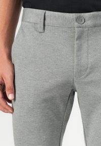 Pantalones texturizados grises con diseño de frente plano, cierre de botón en la cintura y bolsillos laterales. Mano colocada en la zona del muslo.