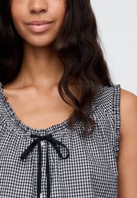 Sort-hvitt gingham-topp med rysjer på ermene, knyting i halsen og knappelukking, laget av en teksturert bomullsstoff.