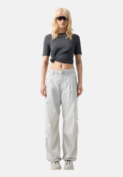Blond kvinde iført sorte solbriller, sort bundet cropped top, løstsiddende hvide bukser med bælte og hvide sneakers, stående mod en ensfarvet baggrund.