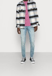 Hombre vistiendo una camiseta rosa, una chaqueta a cuadros en blanco y negro, jeans azul claro y zapatillas negras de caña alta, contra un fondo liso.