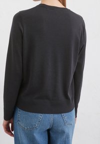 Žena v obyčejném černém svetru s dlouhým rukávem a modrých džínách, zobrazená zezadu na bílém pozadí.