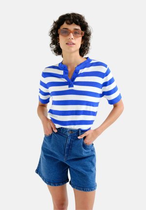 Mujer con cabello rizado que lleva gafas de sol rectangulares, camisa de rayas azules y blancas y pantalones cortos vaqueros de talle alto, manos en los bolsillos.