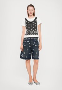 Zwarte bloemenkant overlay op een witte t-shirt, gecombineerd met donkere denim shorts met geborduurde bloemen. Lichtgrijze platte schoenen.