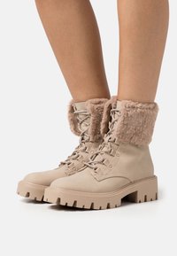 Bottes beiges à lacets avec une semelle épaisse et un revers en fausse fourrure, en cuir lisse avec des oeillets renforcés pour les lacets.