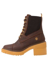 Hnědé kožené kotníkové boty s elastickými bočními panely, žlutými tkaničkami a texturovanou žlutou podešví. Na boku je umístěno logo značky.