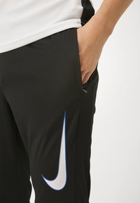 Černé sportovní kalhoty s texturovaným povrchem, s modro-bílým logem Nike na boku a detailem boční kapsy.