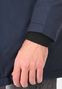 Manica di giacca impermeabile blu navy con polsino nero a coste, caratterizzata da una texture liscia e un design minimalista. La mano poggia su un tessuto testurizzato.
