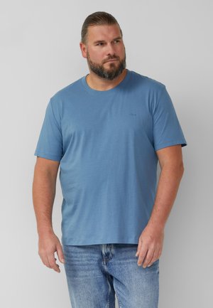 Brodaty mężczyzna z długimi włosami związanymi do tyłu, ubrany w niebieski t-shirt z krótkim rękawem i jasnoniebieskie dżinsy, stojący na tle gładkiego szarego tła.