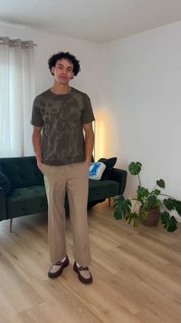 Jonge man met krullend haar, die een donker patroon t-shirt, beige broek en bruine schoenen draagt, staat in de woonkamer met een groene bank en een kamerplant.