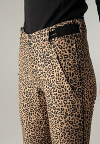 Pantalon imprimé léopard avec fermeture à double bouton noire, poches latérales zippées et bande Velcro noire ajustable, porté avec un haut noir.