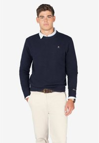 Granatowy sweter dzianinowy z okrągłym dekoltem, teksturową tkaniną i małym haftowanym logo na piersi, w połączeniu z jasnymi spodniami.