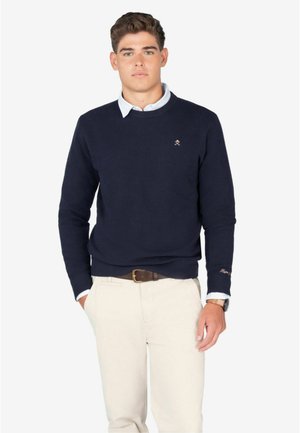 Jersey de punto azul marino con cuello redondo, tejido texturizado y pequeño logo bordado en el pecho, combinado con pantalones de color claro.