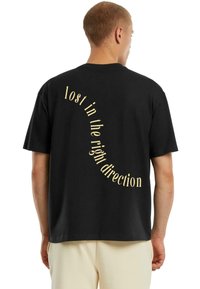 Muž v čiernom tričku s béžovým zakriveným nápisom na chrbte "stratený v správnom smere", skombinovaný s svetlými nohavicami.