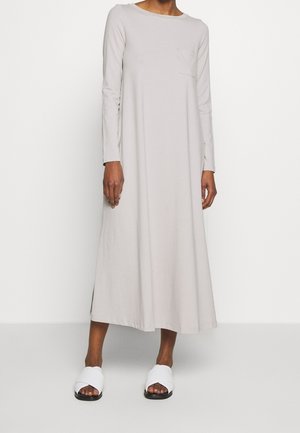 Persona che indossa un abito lungo grigio chiaro a maniche lunghe con una tasca sul petto e sandali bianchi aperti davanti, in piedi davanti a uno sfondo semplice.